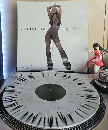 A White W/ Black Splatter vinyl record sits on a turntable. Behind the turntable, a vinyl album outer sleeve is displayed. The front cover shows Christina Aguilera posing with her hair covering her chest. 

To the right of the album cover is an anime figure of Yuki Morikawa singing in to a microphone and holding her arm out. 