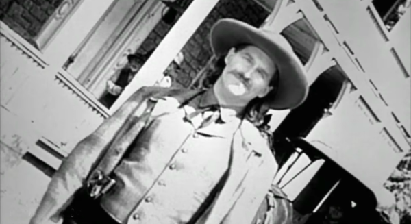 A black and white image of a man with long hair and a cowboy hat. He has a gun on his hip
