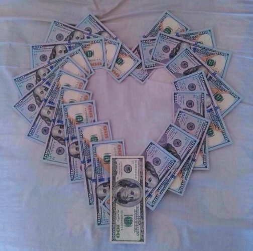 $100 US bills arranged in a heart shape on a wrinkled bedsheet. 
