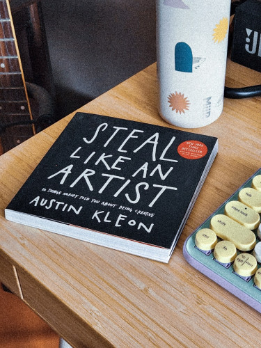 The book called "Steal Like and Artist" by Austin Kleon on a wooden desk. Surrounding it are a water tumbler with cute prints and a mechanical keyboard in pastel hues.