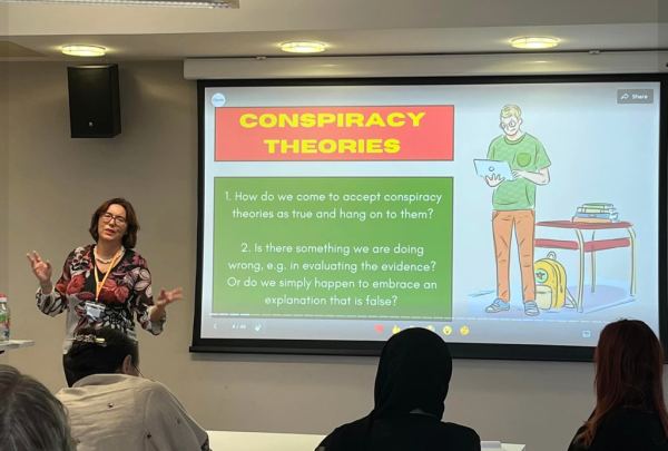Lisa presents at the SAPERE P4C conference. In the background the slides address the key questions in the literature on conspiracy beliefs.