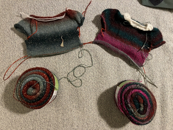 Two tiny jumpers attached to the same long circular needle. To the left reds and greens with gold glitter. To the right reds, pinks and greens with green glitter and a white collar. The colour changing yarn cakes lie below. 