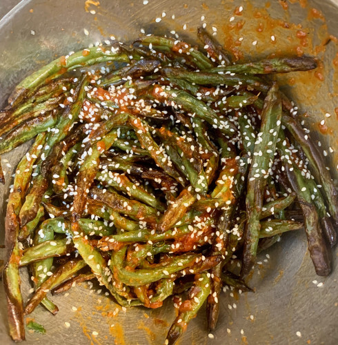 A stainless steel bowl that holds a large handful of roasted green beans, somewhat shriveled and a dark green, with some red sauce and sprinkled with white sesame seeds.
