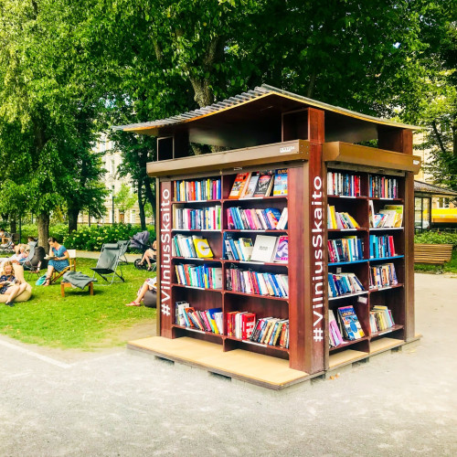 A photo of the Vilnius Skaito public open-air bookshelf in Vilnius, Lithuania, with people sitting on the nearby grass reading. The wooden structure resembles a four-side bookshelf with the hashtag #VilniusSkaito marked on each side and a slightly bent roof.

© Adobe Stock Photographer: Evaldas 

