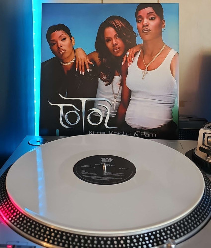 A white vinyl record sits on a turntable. Behind the turntable, a vinyl album outer sleeve is displayed. The front cover shows the 3 members of Total posing together