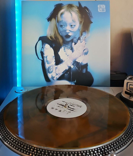 A Orange / Brown Marble [Forest Ash] vinyl record sits on a turntable. Behind the turntable, a vinyl album outer sleeve is displayed. The front cover shows Yeule in a mask and twin tails, She is wearing a sleeveless vest and showing a tattooed right arm, and holding a large cross up to her shoulder