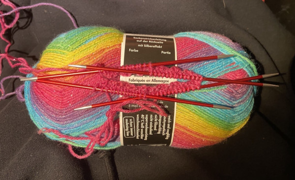 Regenbogenglitzerwollknäuel und zwei Bündchenrunden einer neuen Socke auf vier Stricknadeln 