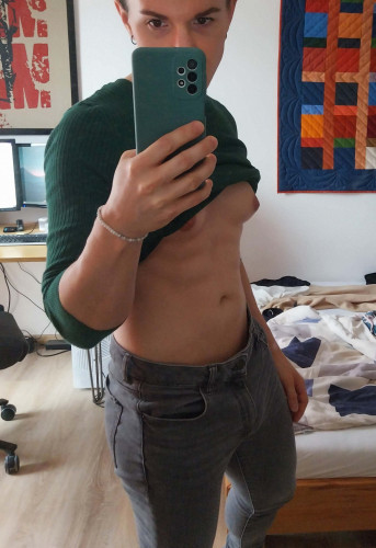 Mirror selfie of a white athletic transfem (me) wearing an emerald green fitting sweatshirt and grey skinny jeans. The sweatshirt is pulled up over her bare breasts exposing her belly and tight abs.