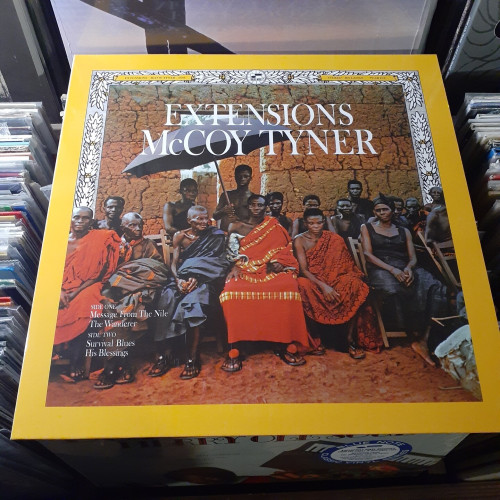 Album cover features a photograph of African village members seated on folding wooden chairs. They are wearing colorful robes, and an earthen wall is behind them. The bright yellow border of the lp and the typeface are in the style of a National Geographic magazine.
