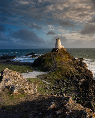 A white painted lighthouse stands on top of a rocky hill. Behind it the sea crashes against the rocks creating lots of white water. The clouds are broken and there is some blue sky visible. The sun is setting to the right of the frame creating shadows and giving the elements in the sun a warm yellow huem
