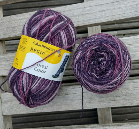 Two balls of yarn in several shades of purple.

On the left the original ball still with the ball and stating it's the brand Regia. On the left the same yarn bit on a hand wound cake.

Everything on a background of weathered wood.