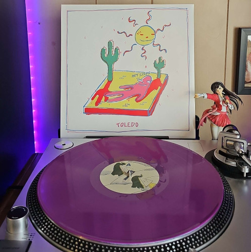 A purple vinyl record sits on a turntable. Behind the turntable, a vinyl album outer sleeve is displayed. The front cover shows a drawing of a blob-like human shape laying in a desert under the sun. 