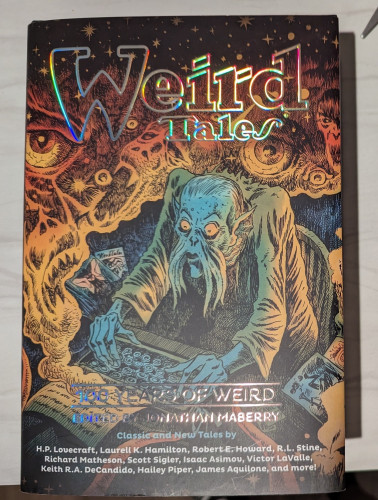 Dust jacket of WEIRD TALES: 100 YEARS OF HORROR that has a strange man typing away on a typewriter while images of various horrors spill out from the pages spooling out of it