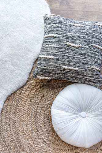 A grey and white square cushion and a white round meditation cushion on a beige woven rug and a fluffy white rug