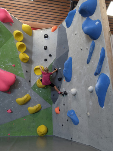 Mädchen mit lila Shirt hängt in etwa in der Mitte einer Boulderwand