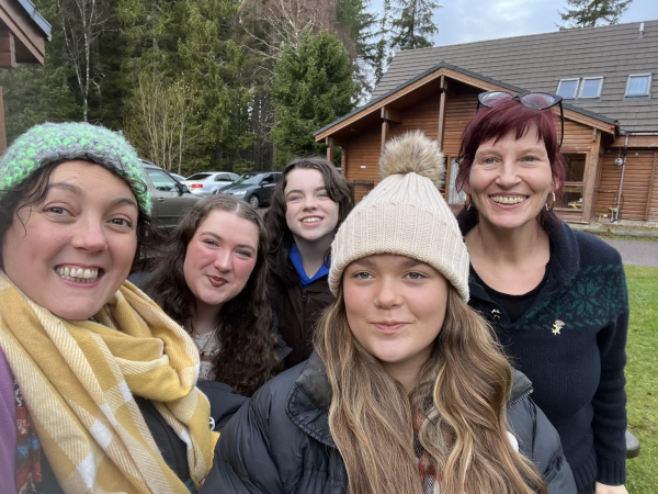 A group of 5 people with two adult women at either school de of 3  young women all smiling to the camera outside a log cabin. 