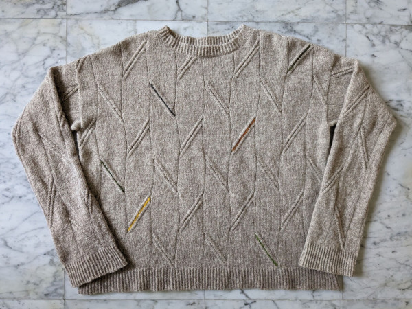 Boxy handknit sweater in a warm grey colour, laid flat. The sweater has a subtle diagonal cable pattern and pops of accent colours in the cable.