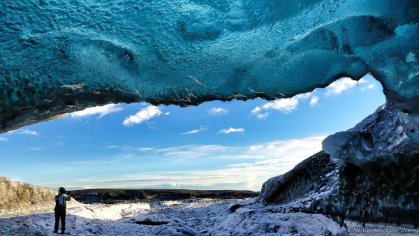 This shot was taken from just inside the mouth of an ice cave, looking outwards. The sky fills the centre of the photo; it's blue with some light puffy clouds. A person is standing in the cave entrance, apparently taking a photo. The floor of the cave is covered with well-trodden snow and ice, the side wall on the right of the shot is deep turquoise-blue with snow sitting in the dimples, and the cave roof is overhead. The clear turquoise ice has light shining through it. In places, snow over the glacier surface is blocking the light and producing darker patches. The light is distorted by ice thickness, and there are a myriad shades of blue and cyan. The whole ceiling looks like it was made of a precious translucent jewel.