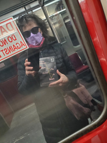 Me, masked, wearing a black winter coat and a pink and purple scarf, holding the novel How to Build a Boat by Elaine Feeney (Biblioasis), reflected in the Toronto Transit Commission (TTC) subway car door, on which a "Danger / Do Not ..." sign is partially visible