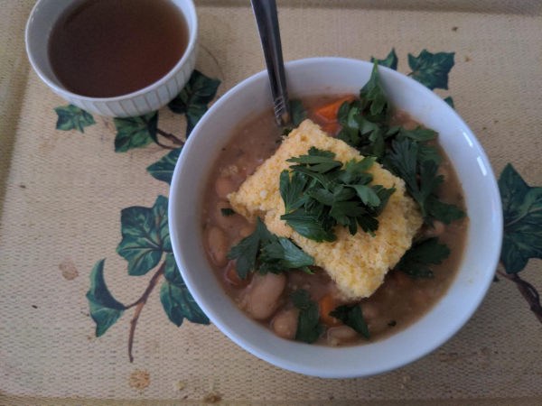 A bowl of white beans with a big chunk of cornbread, some carrot chunks, and some parsley, with a spoon. An Asian-style (handle-less) teacup with black tea. Placed on a dirty yellow metal food tray with leaf pattern.