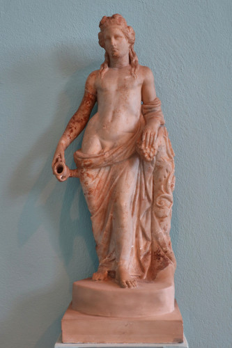 Reddish terracotta statuette of the god Dionysos leaning on a column holding a cluster of grapes in his left hand and a small kantharos cup in his right. He is wearing a grapevine crown and his long hair flows over his shoulders. A himation is wrapped around his legs and left arm, leaving his genitals bare.