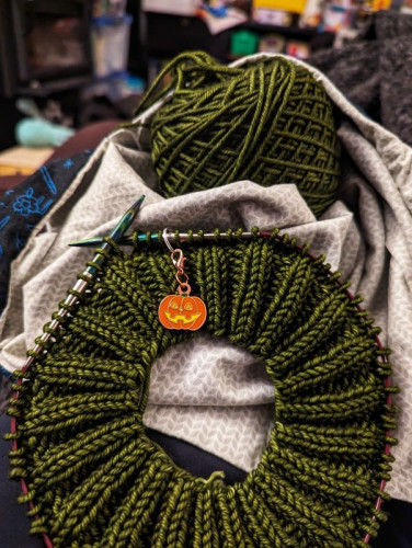 A knitted hat brim in dark green yarn with a Halloween pumpkin stitch marker showing the beginning of the round.