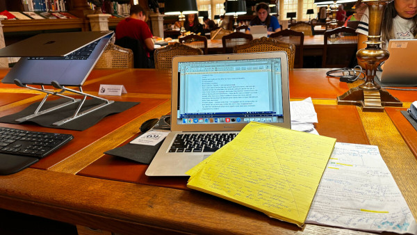 A laptop on a library desk, with papers askew in front and sunglasses behind.