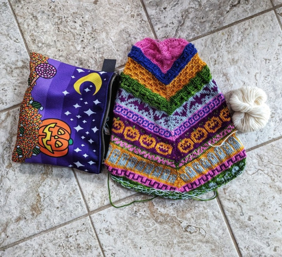 A scarf-style cowl with Halloween motifs on it sits next to a Halloween themed bag with a jack-o-lantern on it. On the other side is a brighter white ball of yarn with the tail over an orange section to show some contrast.