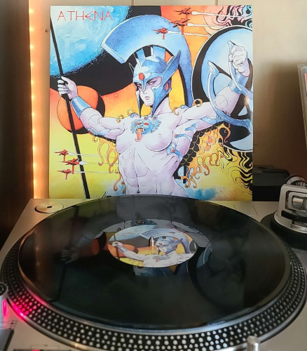 A black vinyl record sits on a turntable. Behind the turntable, a vinyl album outer sleeve is displayed. The front cover shows artwork of a Topless athena in a helmet, and holding a shield and spear. Spaceships are flying around her, and planets in the background. 
