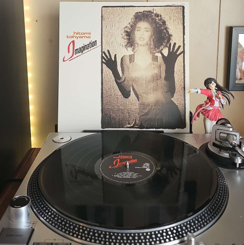 A black vinyl record sits on a turntable. Behind the turntable, a vinyl album outer sleeve is displayed. The front cover shows Hitomi Tohyama behind glass with some type of film on it. Her hands are pressed against it as she looks at the camera. 

To the right of the album cover is an anime figure of Yuki Morikawa singing in to a microphone and holding her arm out. 
