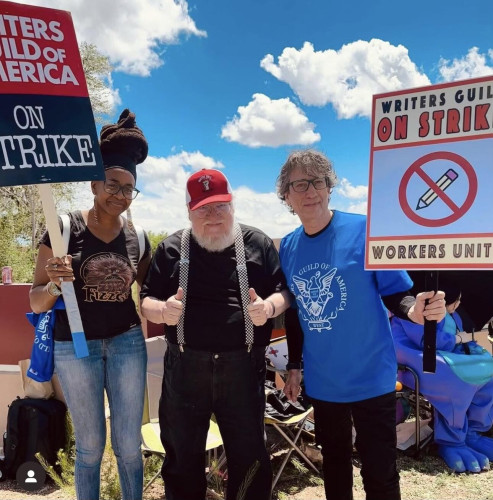Author George R. R. Martin standing with authors Nnedi Okorafor and Neil Gaiman, both of whom are holding up signs for the writer's guild strike.