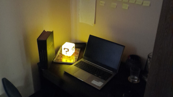 A moody shot of a writer's desk, with a laptop, Post-Its and a glowing lamp in the shape of a Question Block on top of a notebook.