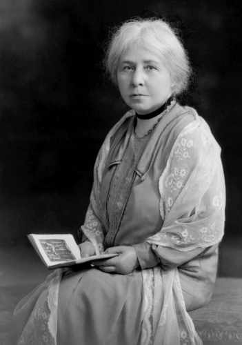 A black and white photograph portrait of Margaret Murray. She sits, gazing at the camera, holding a book which is open, as if she was interrupted while reading.