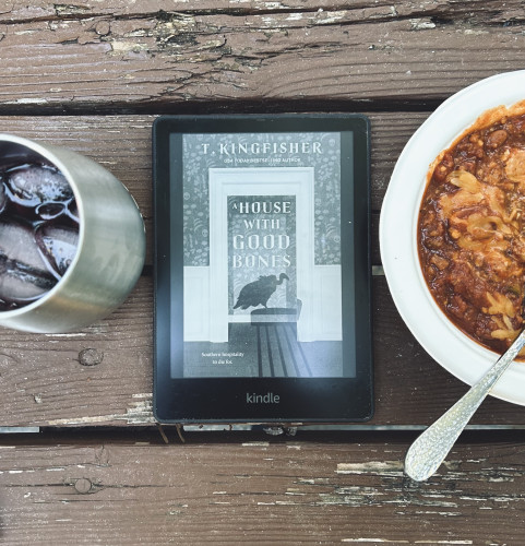 A glass of wine, my Kindle, showing the cover: A House With Hood Bones by T. Kingfisher,  & a bowl of chili.