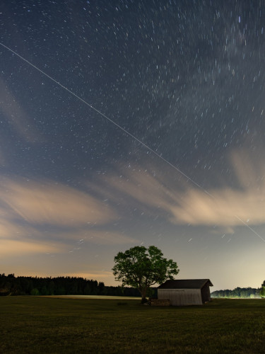longexposure image of the nightsky with the ISS as a long streak from the top left to the bottom right. In the foreground is a wooden barn and big tree on a meadow. The stars are forming short streaks in a circle around the celestial north pole due to the long exposure. some motion blurred clouds are also visible in the sky.