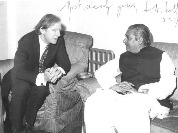 On the right is pictured the first President of Bangladesh sitting on a chair and smiling, looking at Peter Shore (sitting on a separate chair the left) who is talking. 