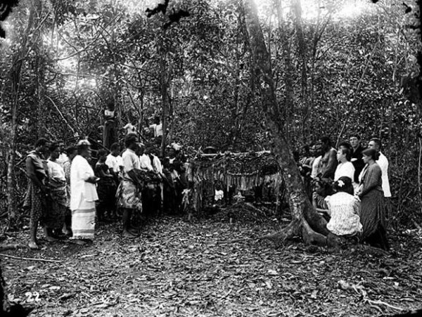 A black-and-white photograph, showing the burial of Robert Louis Stevenson. A number of people stand solemnly in a thickly wooded area around what must be Stevenson's coffin, draped in cloth and leaves.