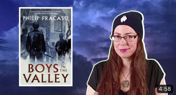 The book cover for Boys in the Valley by Philip Fracassi with a stormy bluish violet background and me (she/her with long brown hair) next to the book wearing a navy beanie with a ghost on it and a black shirt.