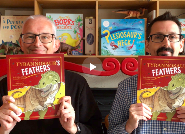 Still from a video of Jonathan and Adam each holding up a copy of The Tyrannosaur's Feathers book with other books, and dinosaur toys, on shelves in the backgroud.