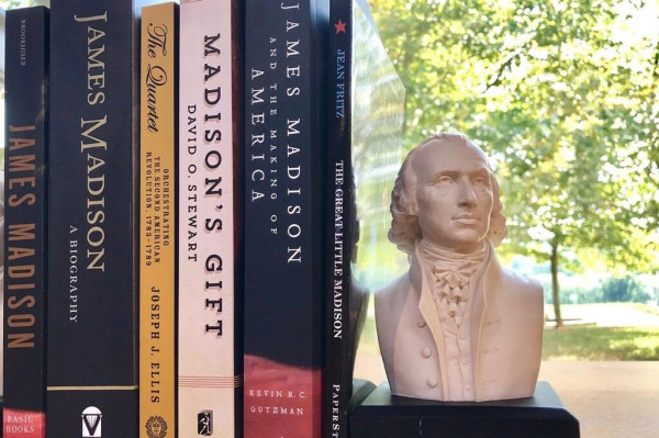 A bust of James Madison sits next to a line of books with titles all centered on the life of James Madison. Green trees and grass can be seen behind the bust, through the window to outside. 
