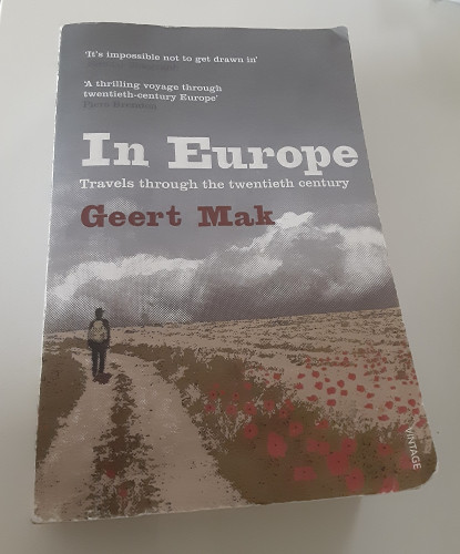 Image shows a paperback copy of Geert Mak's book In Europe: a man facing away from the viewer, backpack on his back and standing on a path to the left of brown fields with red flowers, with most of the cover taken up by gray-blue sky and big low-horizon clouds above the field.