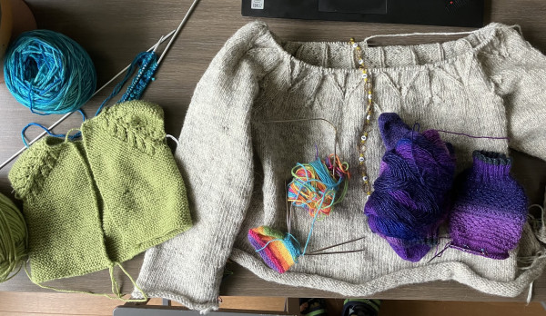A bunch of wool and knitting on a table. Left, a turquoise ball of yarn with little more than a cast-on on long double pointed needles. On top of the needles, a light green garter stitch baby cardigan sans sleeves, with lace detail around the raglan. Next to that a grey sweater with rolling hems, knitted up to part of the yoke. On top of it a rainbow baby booty and a ombré purple glitter sock. 