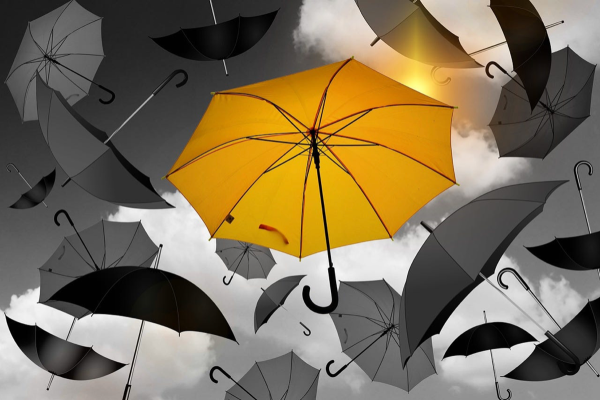 A single golden yellow umbrella falling in a grayscale sky filled with falling grayscale umbrellas.