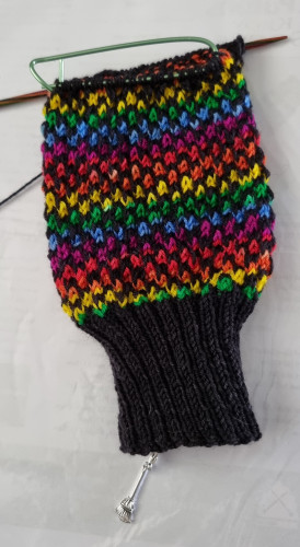 Half a knitted sock. Cuff is black and body of sock is knitted using rainbow stripes wool. Two stitches if colour alternating with two of black.