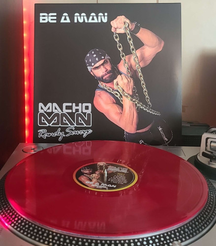 A red vinyl record sits on a turntable. Behind the turntable, a vinyl album outer sleeve is displayed. The front cover shows Macho Man Randy Savage wearing a large and super long gold chain and holding it up with his fists. 
