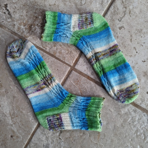 A pair of (almost) matching socks with stripes of green, blue and white with a purple-ish variegated section. They have a fancy cable pattern on them that is mirrored on each sock. 