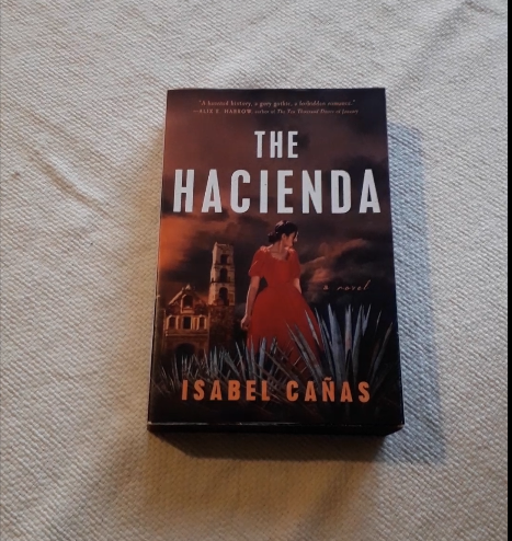 In the cover of The Hacienda A woman in a red dress stands between magueyes and a the tower of a old church. The ominous sky is red and covered y gray clouds.