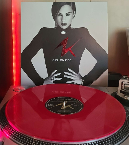 A red vinyl record sits on a turntable. Behind the turntable, a vinyl album outer sleeve is displayed. The front cover shows Alicia Keys posing with her hands on her hips as she looks at the camera. 