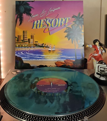 A Transparent Sea Green vinyl record sits on a turntable. Behind the turntable, a vinyl album outer sleeve is displayed. The front cover shows a beach with buildings on the other side of the short. Sailboats are in the water . 

To the right of the album cover is an anime figure of Yuki Morikawa singing in to a microphone and holding her arm out. 