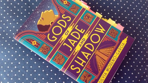 The cover of Gods of Jade and Shadow shows a pyramid at the bottom, the tittle surrounded by Mayan iconography and the profile of a young woman, her face tilted as if looking to the stars, her eyes closed. 
